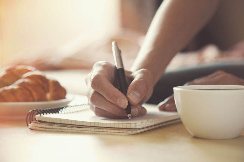 female hands with pen writing on notebook with morning coffee and croissant